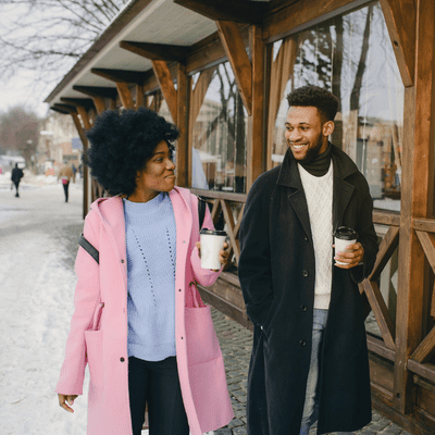 girl in pink jacket and guy in black jacket walking together with white coffee cups in their hands .date night for married couples ideas