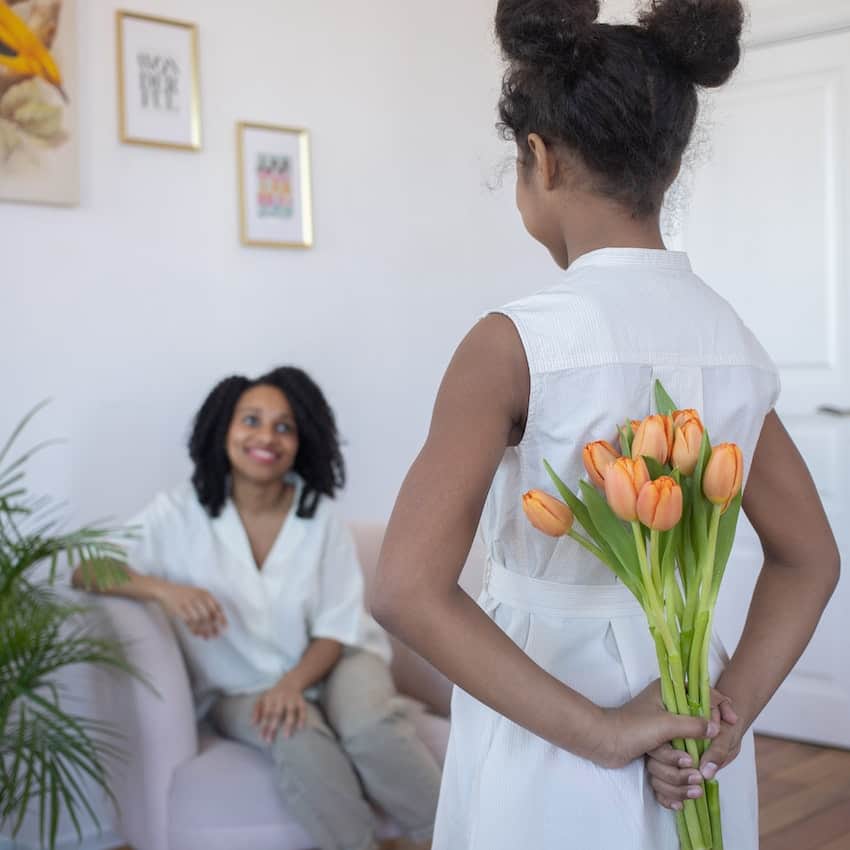 Photo of a girl hiding tulips behind her back for her mom. Things to do for mothers day at home. 